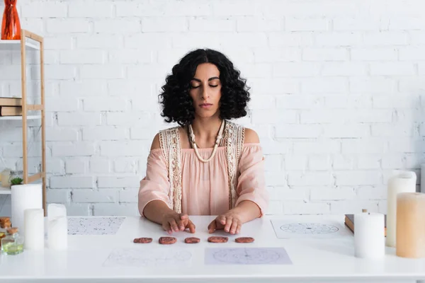 Young astrologer with closed eyes sitting near clay runes and natal charts during spiritual session — Photo de stock