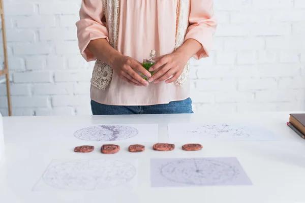 Partial view of fortune teller holding vial of essential oil near clay runes and astrological maps — Stock Photo