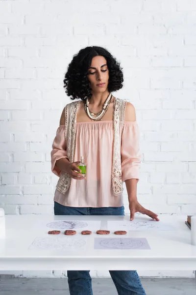 Brunette astrologist with bottle of essential oil standing near clay runes and constellation maps — Fotografia de Stock