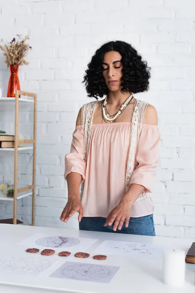 Brunette fortune teller with closed eyes standing near clay runes and cosmic charts during spiritual session — Stock Photo