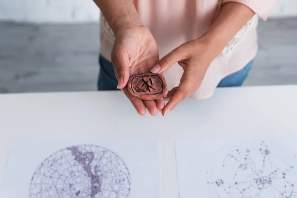 Partial view of astrologer holding clay rune near blurred celestial charts — Stock Photo