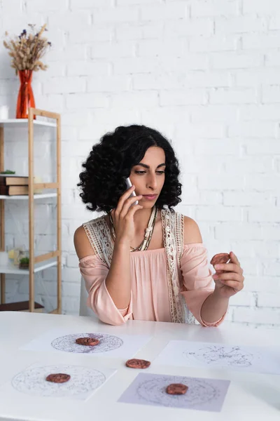 Young astrologer looking at clay rune and talking on mobile phone at home — Fotografia de Stock