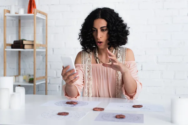 Brunette soothsayer gesturing during video call on smartphone near clay runes and stars charts — Photo de stock