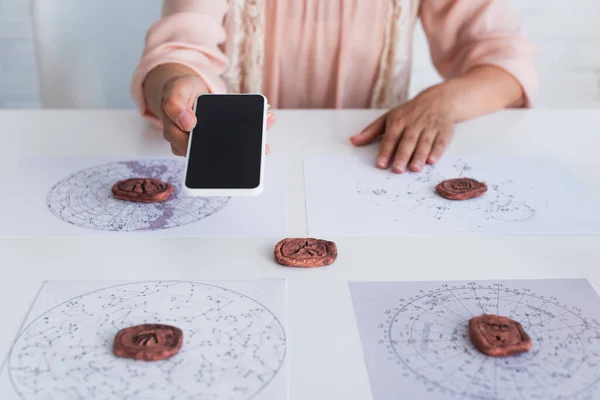 Cropped view of oracle holding mobile phone with blank screen near celestial maps and clay runes — Stock Photo