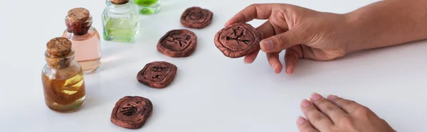 Cropped view of predictor holding clay rune near bottles with essential oils, banner - foto de stock