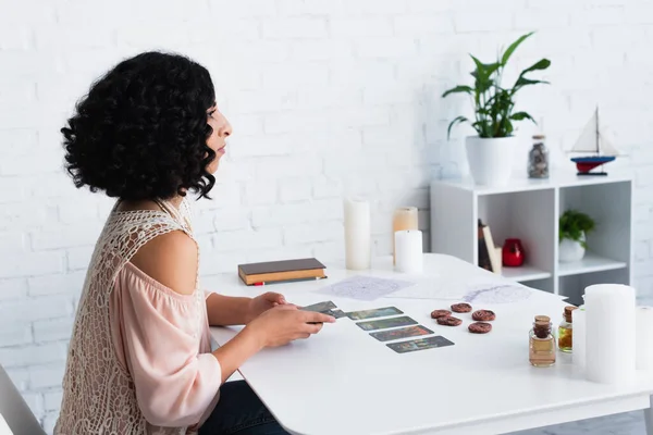 KYIV, UKRAINE - JUNE 29, 2022: side view of brunette soothsayer with tarot cards near clay runes and essential oils — Stock Photo