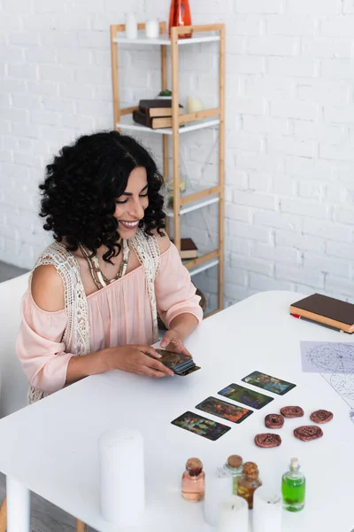 KYIV, UKRAINE - JUNE 29, 2022: smiling astrologer holding tarot cards near clay runes and bottles with essential oils — стокове фото