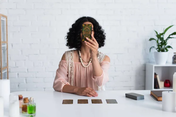 KYIV, UKRAINE - JUNE 29, 2022: brunette astrologer obscuring face with tarot card during spiritual session at home — Foto stock