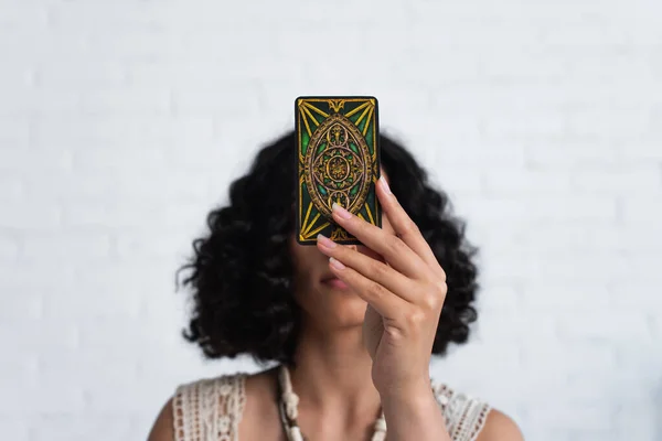 KYIV, UKRAINE - JUNE 29, 2022: young brunette oracle obscuring face with tarot card during spiritual session — Foto stock
