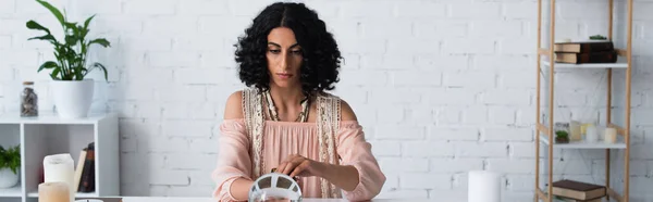 Young astrologer sitting near crystal ball and candles during prediction session, banner — Stock Photo