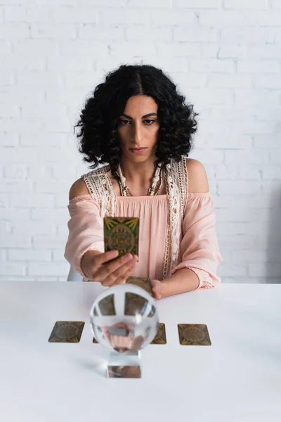 KYIV, UKRAINE - JUNE 29, 2022: brunette woman holding tarot cards near blurred crystal ball at home — Photo de stock