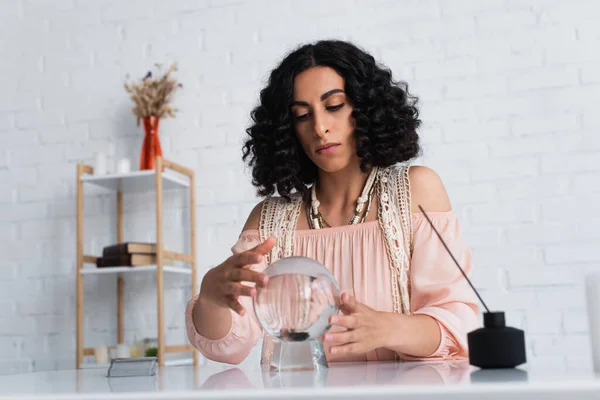 Brunette soothsayer holding hands near crystal ball and diffuser with aroma stick — Stock Photo