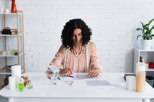 Smiling astrologer looking at celestial charts near crystal ball and bottles with essential oils — Stock Photo