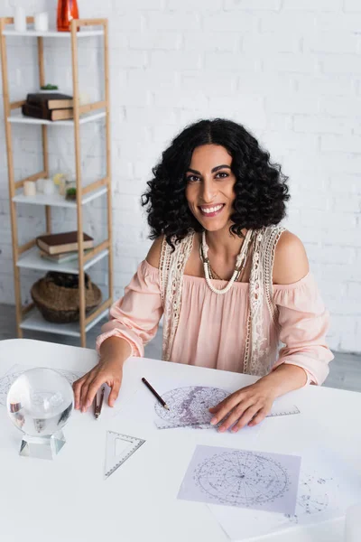 Brunette astrologer smiling at camera near crystal ball and star charts — Foto stock