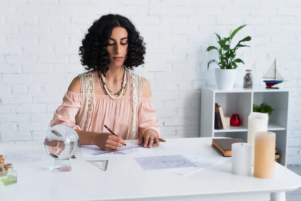 Young astrologer drawing natal charts near crystal ball and candles at home — Fotografia de Stock