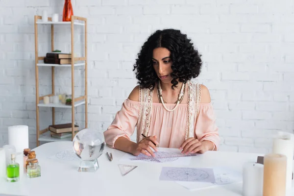 Young soothsayer drawing celestial charts near crystal ball and bottles with essential oils — Stock Photo