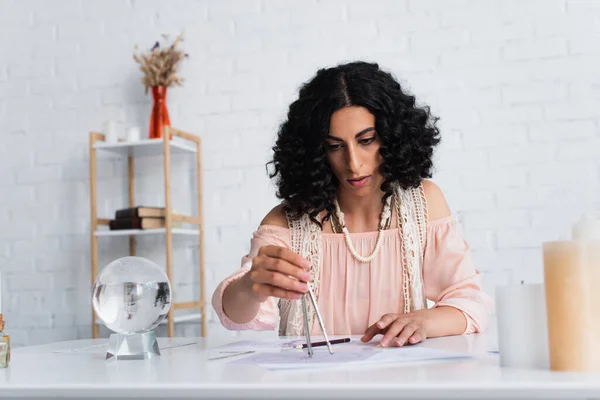 Brunette astrologer drawing star charts near crystal ball at home — Fotografia de Stock