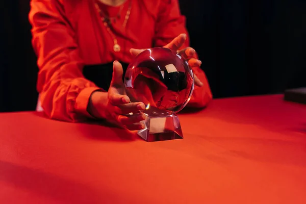 Partial view of fortune teller near crystal ball on red table isolated on black - foto de stock