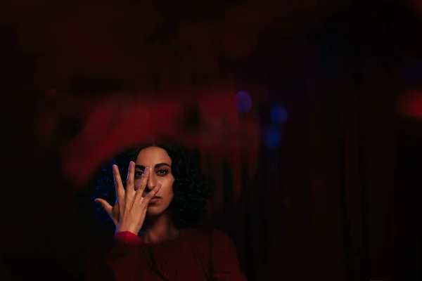 Portrait of brunette soothsayer obscuring face with hand and looking at camera on dark background - foto de stock