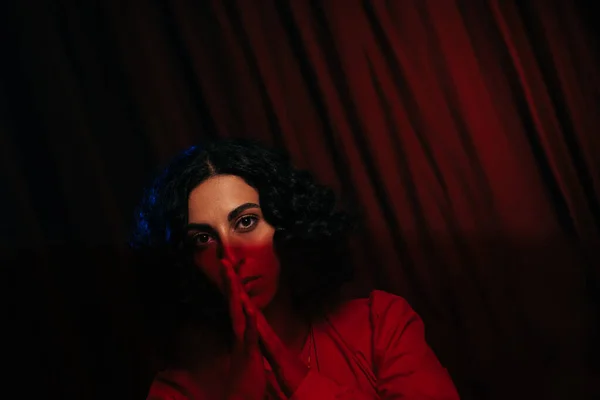 Brunette oracle with folded hands near face looking at camera on dark background with red drape - foto de stock
