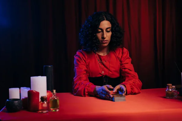 Brunette medium sitting near tarot cards and candles on dark background with red drape - foto de stock