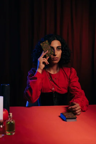 KYIV, UKRAINE - JUNE 29, 2022: brunette fortune teller obscuring face with tarot card on dark background with red drape — Stock Photo