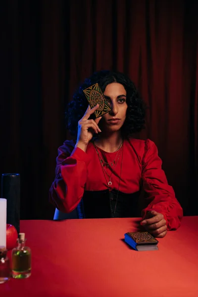 KYIV, UKRAINE - JUNE 29, 2022: brunette fortune teller obscuring face with tarot card on dark background with red drape — стокове фото