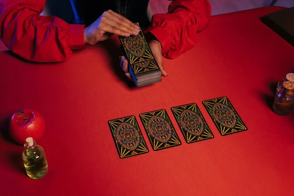 KYIV, UKRAINE - JUNE 29, 2022: partial view of oracle laying out tarot cards on red table near essential oils - foto de stock