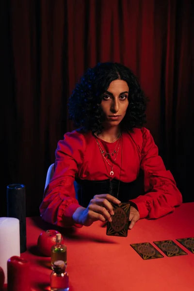 KYIV, UKRAINE - JUNE 29, 2022: young brunette medium with tarot card looking at camera on dark background with red drape — Stock Photo