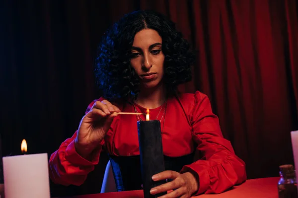 Young oracle lighting aroma stick near burning candle on dark background with red drape — Photo de stock