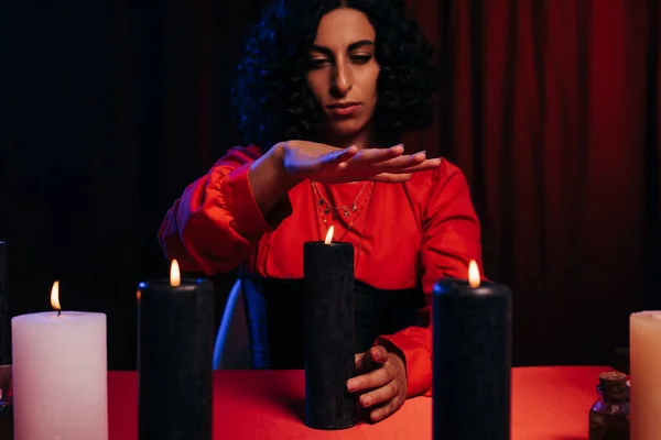 Brunette medium holding hand over burning candle on dark background with red drape — Stock Photo
