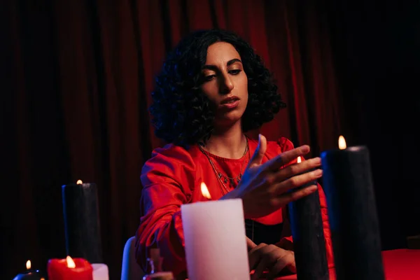 Young fortune teller sitting near burning candles on dark background with red drape — Stock Photo