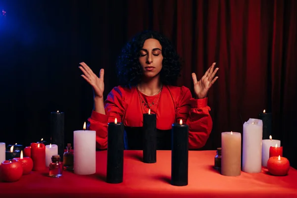 Young oracle with closed eyes during spiritual session near burning candles on dark background with red drape — Foto stock