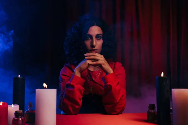 Young medium sitting near burning candles on dark background with blue smoke and red drape — Stock Photo