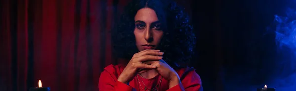 Fortune teller looking at camera on dark background with red drape and blue smoke, banner — Stock Photo