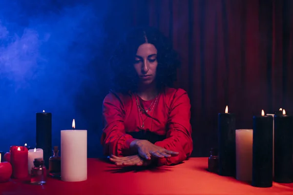 Fortune teller sitting during magic session near burning candles on dark background with blue smoke — Stock Photo