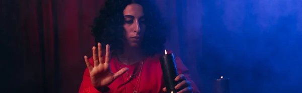 Young oracle holding burning candle during spiritual session in darkness with blue smoke, banner - foto de stock
