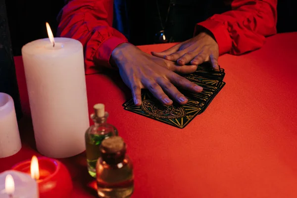 KYIV, UKRAINE - JUNE 29, 2022: partial view of medium with tarot cards near burning candles and essential oils isolated on black — стоковое фото