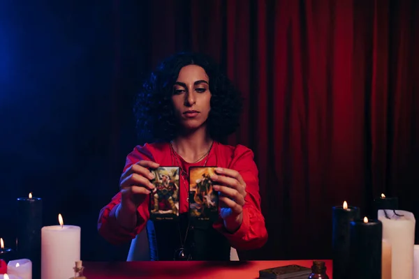 KYIV, UKRAINE - JUNE 29, 2022: brunette oracle holding tarot cards on dark background with red drape — Stock Photo