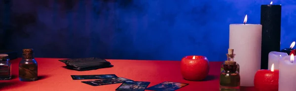 KYIV, UKRAINE - JUNE 29, 2022: essential oils and tarot cards near burning candles on red table near blue smoke on dark background, banner - foto de stock