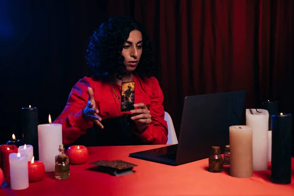 KYIV, UKRAINE - JUNE 29, 2022: brunette oracle holding tarot card during online esoteric session on laptop on black background with red drape — Stockfoto