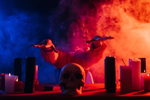 Oracle with closed eyes holding hands above skull during spiritual session in darkness near colorful smoke - foto de stock