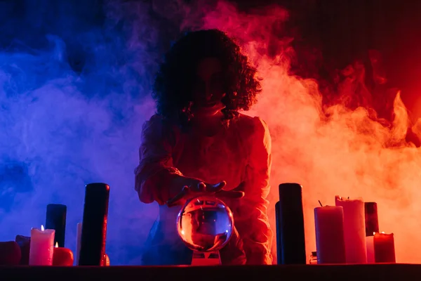 Young fortune teller near crystal ball on dark background with colorful smoke — Stock Photo