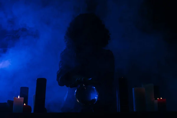 Fortune teller holding hand above crystal ball near burning candles in darkness with blue smoke — Stock Photo