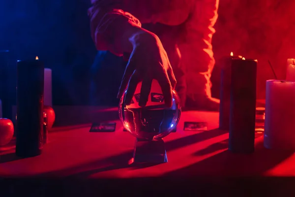 Cropped view of medium touching crystal ball near burning candles on dark background with smoke — Stock Photo