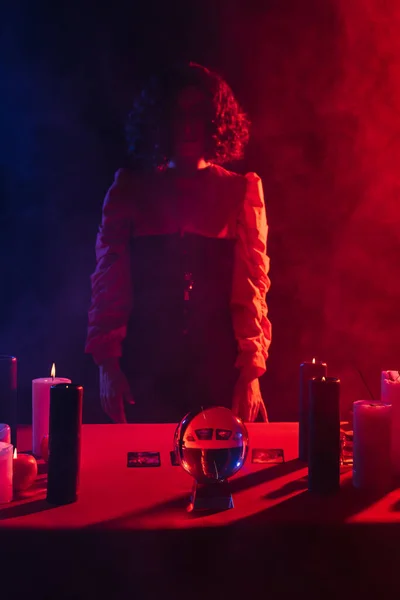 Young fortune teller standing near red table with candles and crystal ball on black background with smoke — Foto stock