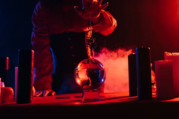 Cropped view of medium holding amulet over crystal ball on black background with smoke — Stock Photo