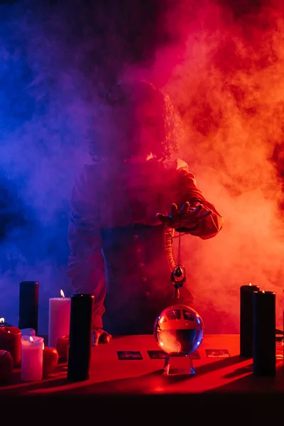 Fortune teller holding amulet near crystal ball and candles near colorful smoke on foreground — Stock Photo