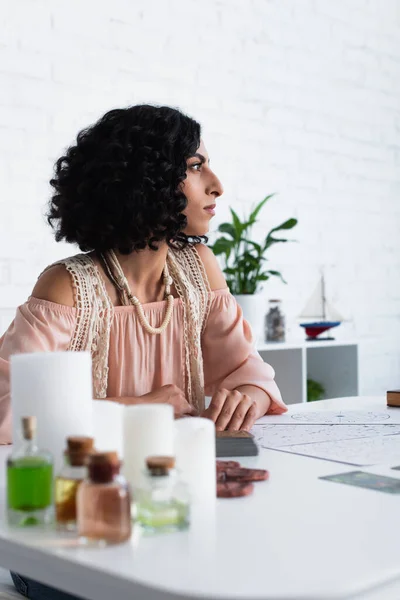 Young astrologist looking away near candles and bottles with essential oils — стоковое фото