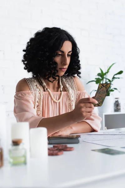 KYIV, UKRAINE - JUNE 29, 2022: young oracle looking at tarot card near blurred candles — стокове фото